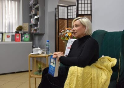 Na zdjęciu widoczna jest czytelnia Biblioteki w Zelowie podczas spotkania autorskiego z pisarką Lidia Liszewska. Gość wydarzenia siedzi na fotelu i prezentuje uczestnikom książkę swojego autorstwa.